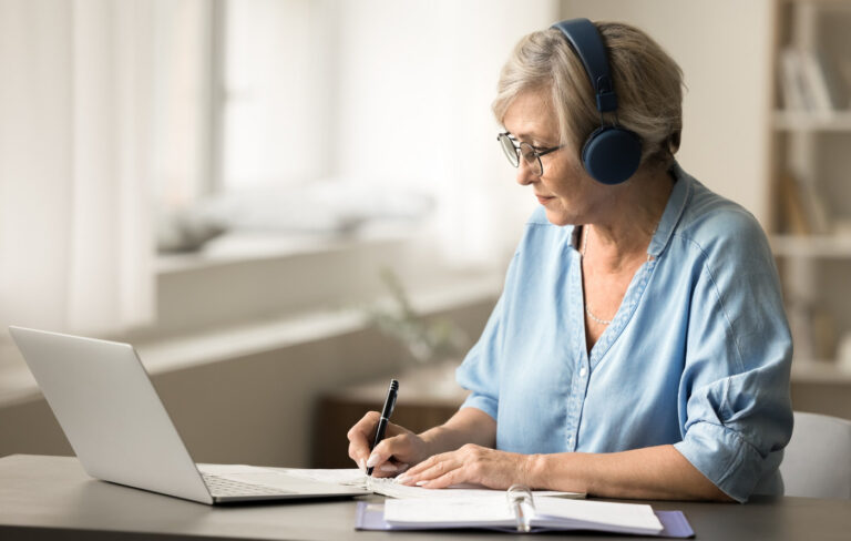 Focused senior woman wearing headphones, looking at a laptop and writing notes
